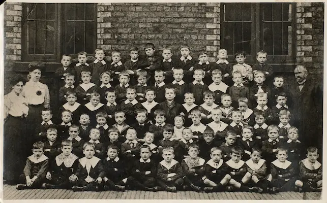Archival group photograph of a large class of schoolboys posed outdoors with two female teachers and a male teacher against a brick school building.