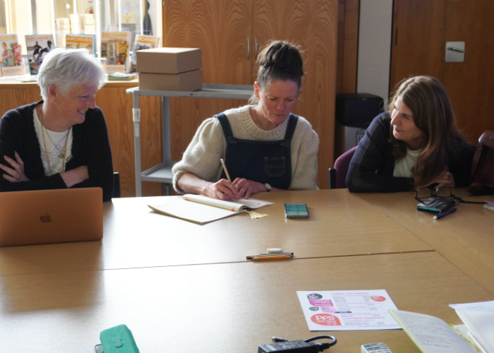 Three women sitting around a table in an archive room, collaborating whilst one writes in a notebook next to a laptop and smartphone.