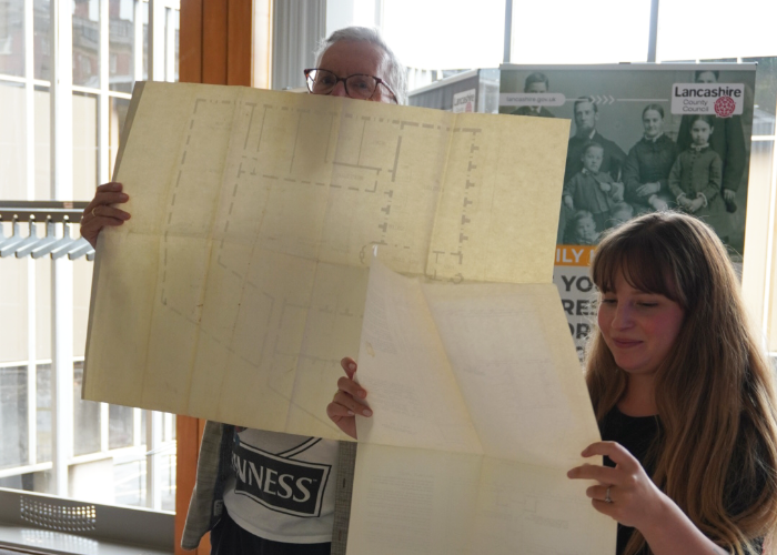 Two people holding up large, unfolded historic architectural plans and documents in front of a Lancashire County Council banner.