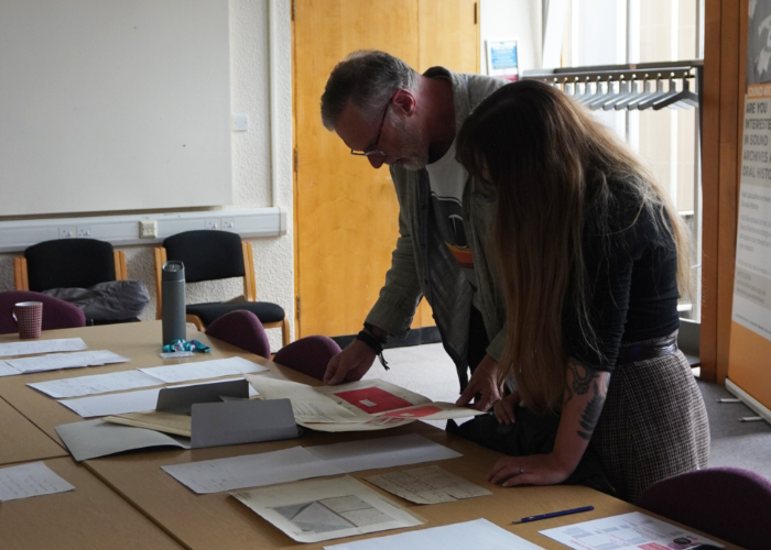 Two researchers leaning over a table to study old documents and files in an archive room.