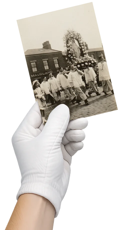 A white-gloved hand holding an old black-and-white photograph against a black background. The vintage photo shows a religious procession on a cobblestone street, with people in white robes carrying a large, flower-adorned statue of the Virgin Mary. A brick building with a “MAUDLAND” sign is visible in the background.