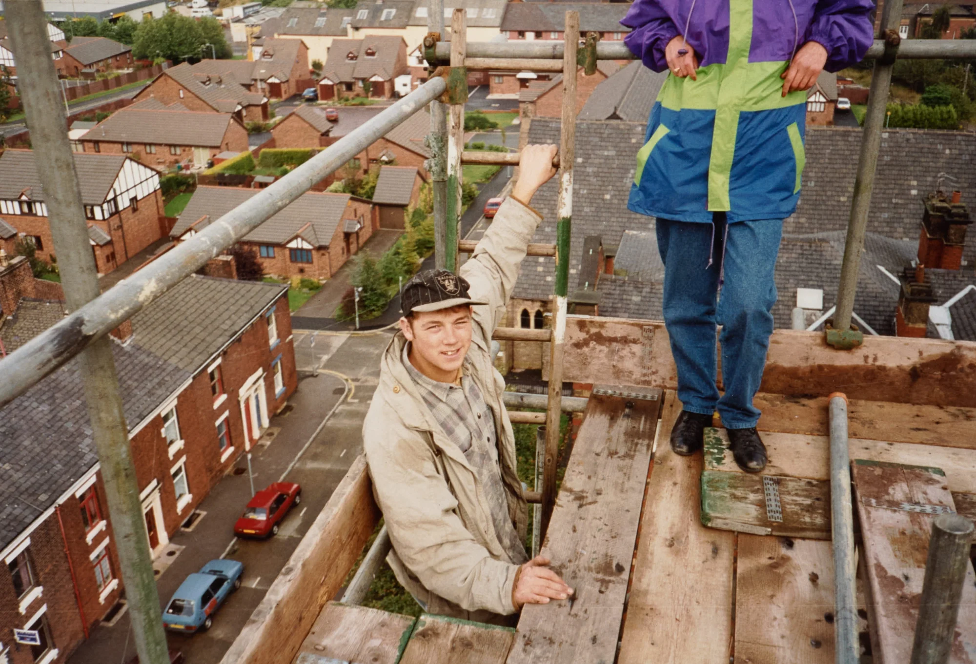 Photograph taken from scaffolding near the top of St Walburge's tower, looking down over the terraced streets and rooftops of the parish. A young workman climbs onto the boarded platform; a second worker stands beside him.