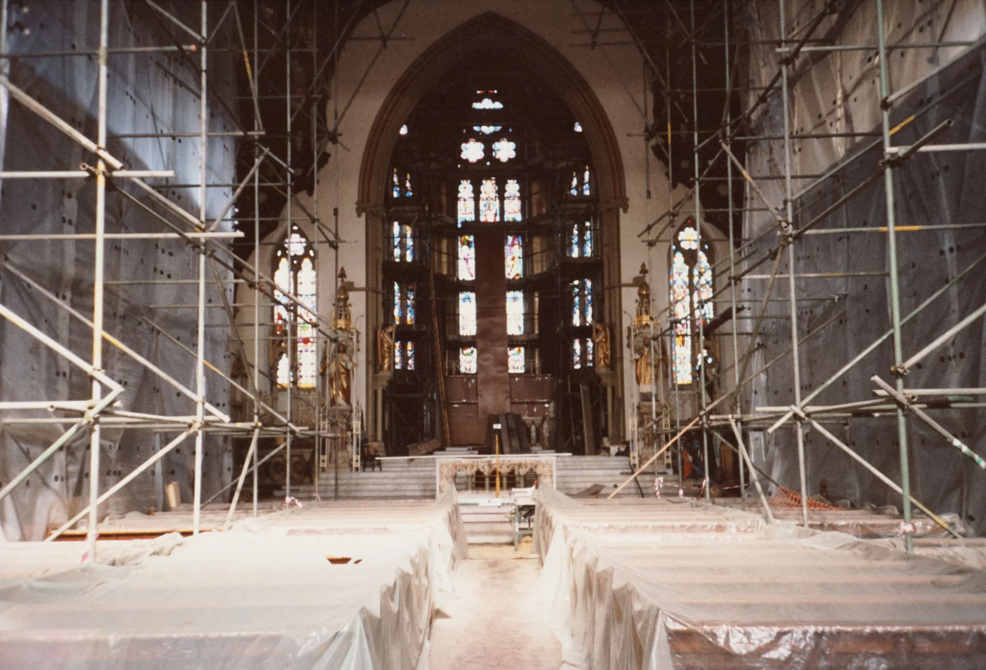 Photograph looking down the nave of St Walburge's during restoration, the pews sheeted in dust covers and scaffolding rising the full height of the walls either side. At the far end, the great east window and gilded statues remain visible above the empty sanctuary.