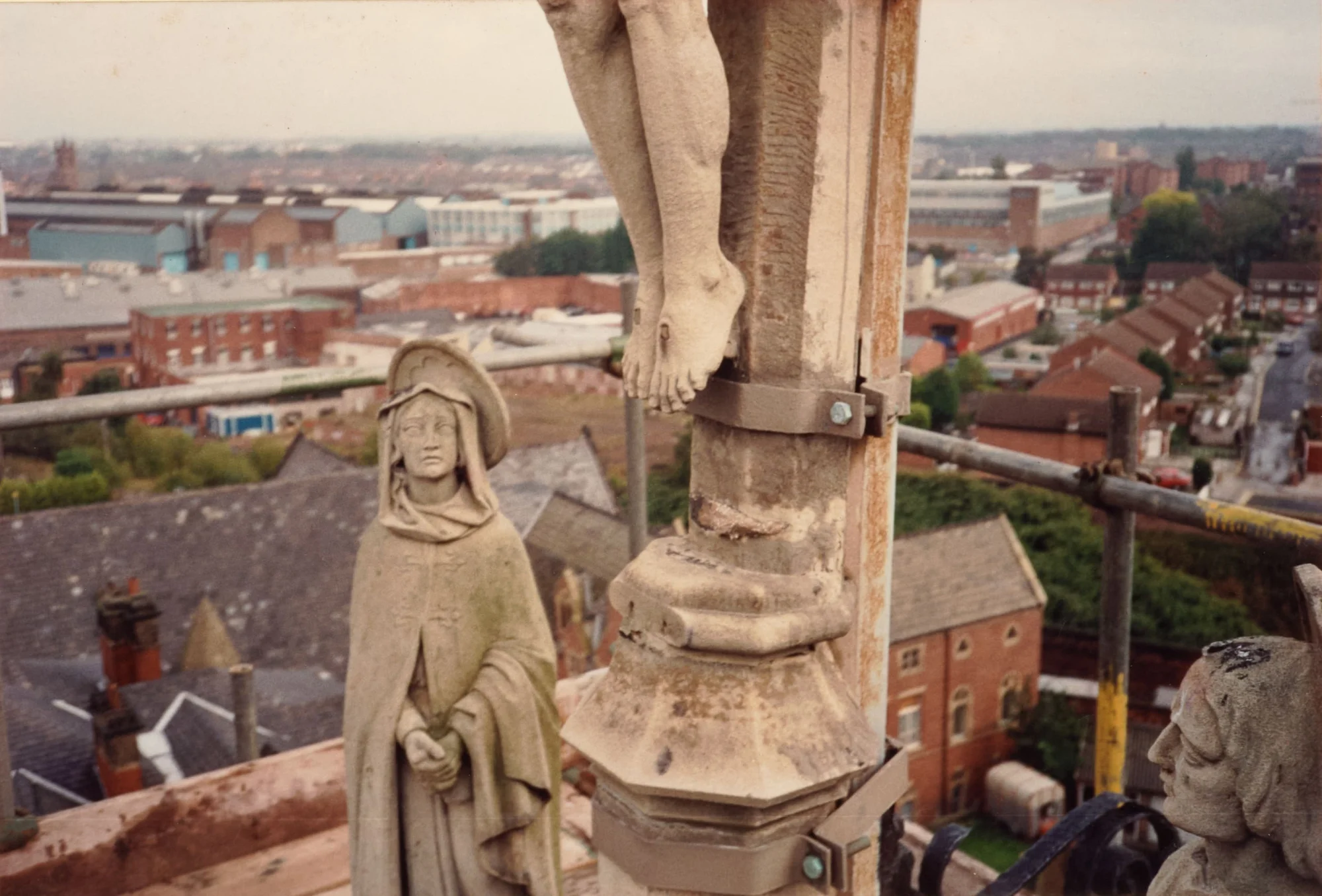 Photograph taken at height beside the Calvary above the west front entrance doors of St Walburge's: the feet of the crucified Christ above, with statues of Our Lady and St John standing either side. Scaffolding and Preston's rooftops stretch out beyond.