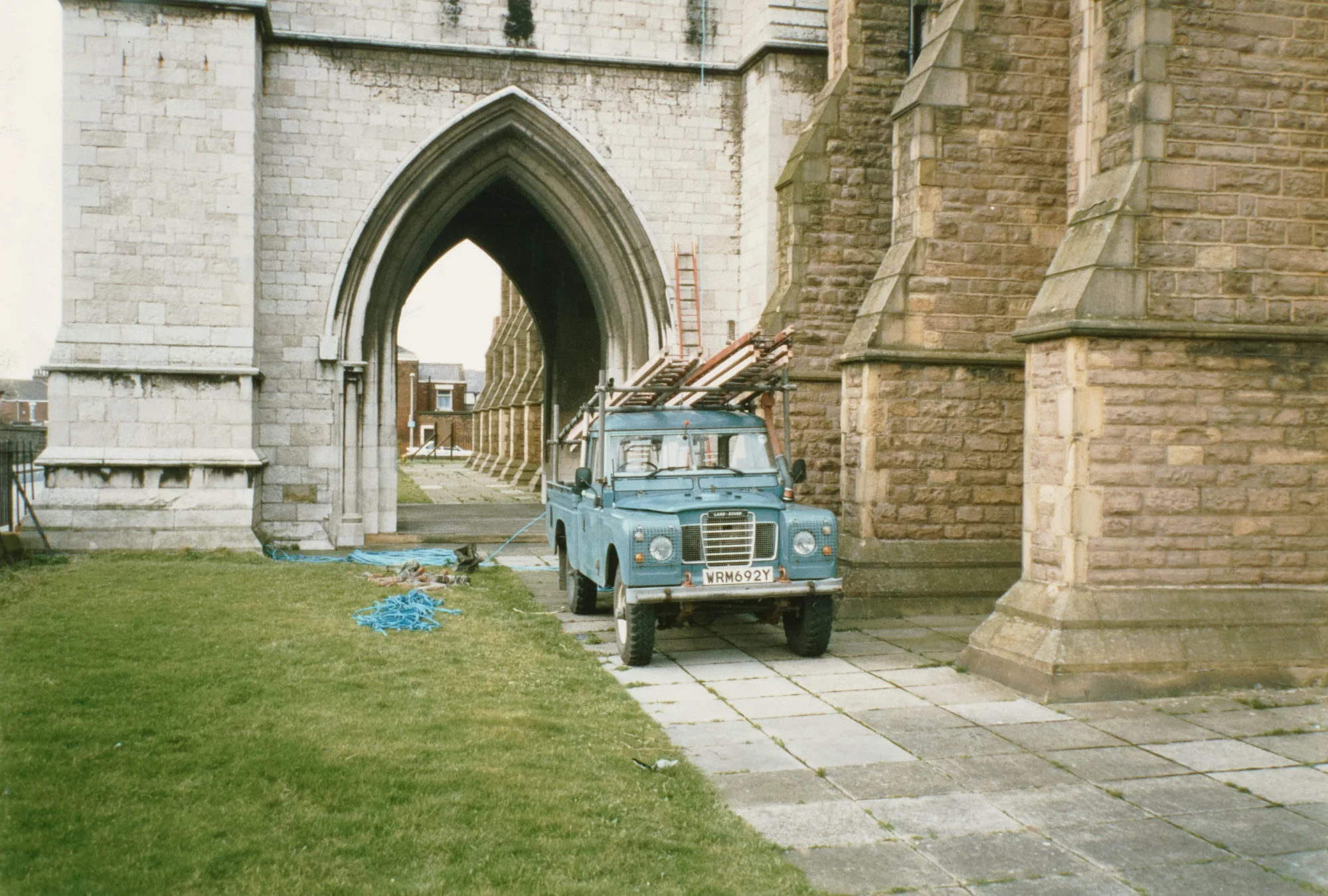 Photograph of a blue Land Rover loaded with ladders, parked on the paving beneath St Walburge's arched west porch, coiled ropes lying on the grass beside it. The pointed archway frames a further view of the covered walkway and houses beyond.