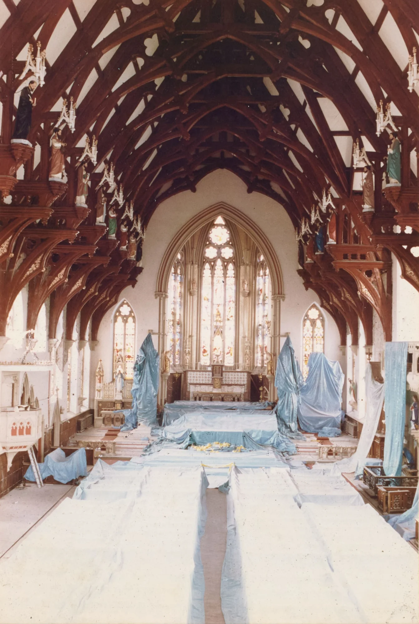 Photograph looking down the nave of St Walburge's toward the sanctuary, the great timber roof arching overhead and statues of saints standing on brackets along both sides. The altar and furnishings are wrapped in dust sheets, with the pews beneath covered from end to end.