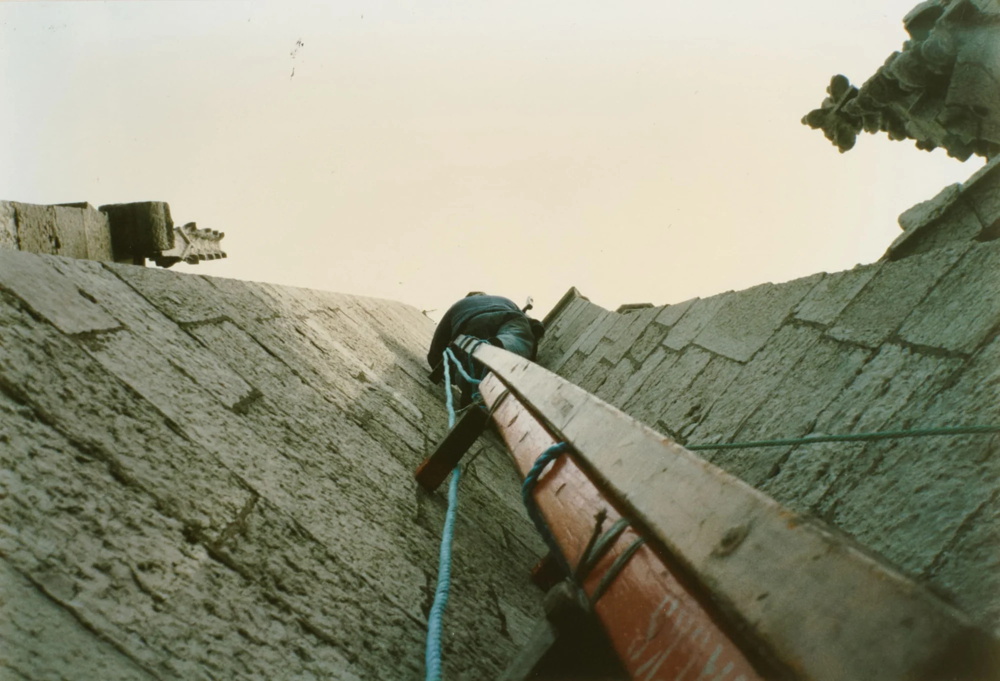 Photograph looking up the steep slate roof ridge of St Walburge's, a workman climbing near the top secured by rope, a ladder laid against the slope beside him. Carved stone ornaments project from the roofline against the sky.