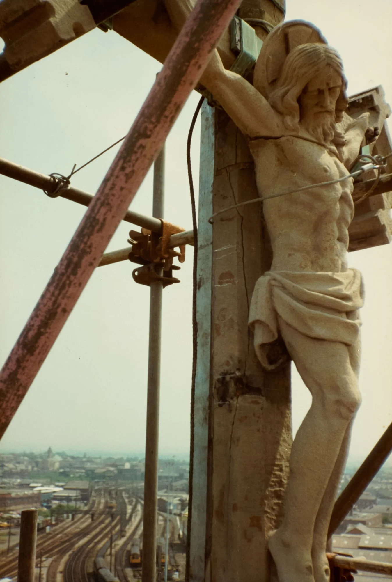 Photograph taken close beside the crucifix of St Walburge's Calvary, secured against its cross with a temporary wire during restoration, scaffolding poles crossing the frame. Preston's railway lines and rooftops visible in the distance.