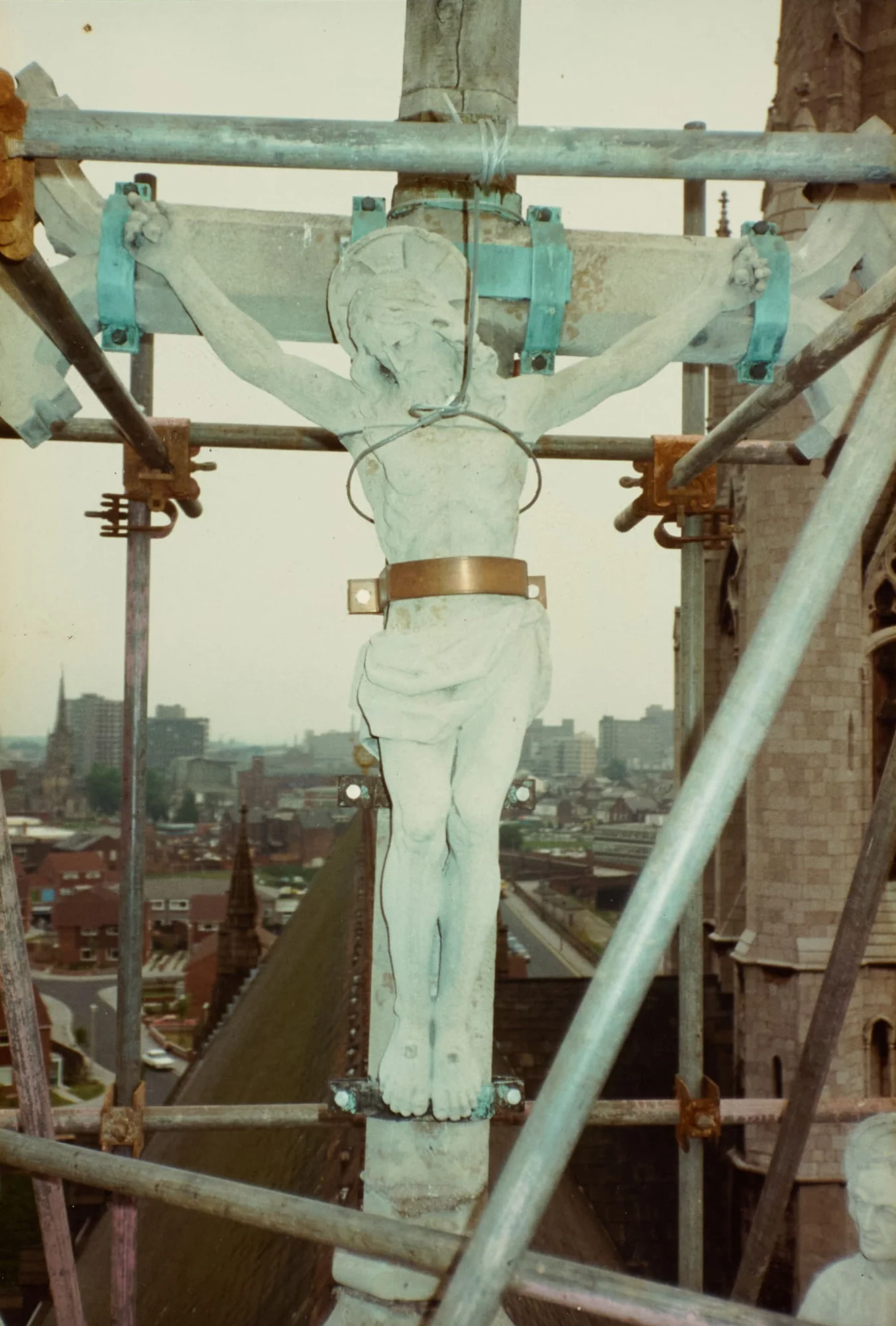 Photograph of the crucifix of St Walburge's Calvary, seen close and full-length, fixed to the cross with metal brackets and bound at the waist as scaffolding is erected around it. Preston's rooftops and skyline stretch out below.