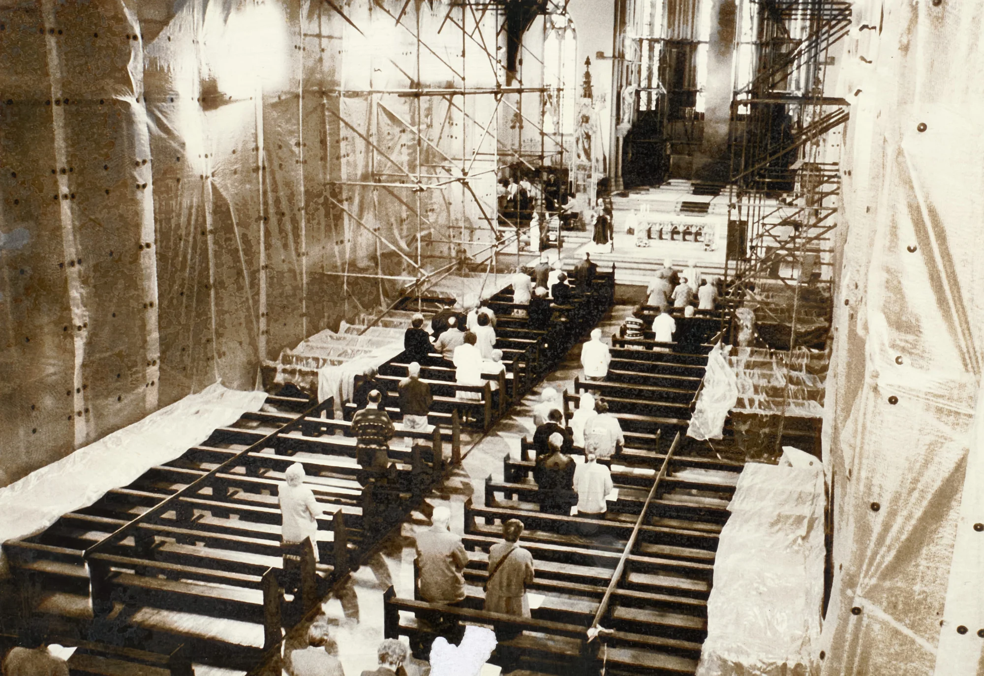Photograph looking down the nave of St Walburge's during restoration, the congregation seated in the pews for Mass while scaffolding rises to the roof on either side and sheeting hangs from the walls. The priest stands on the sanctuary.