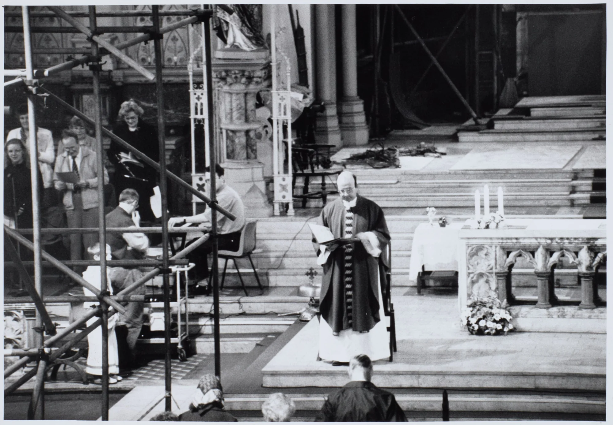 Photograph taken during Mass at St Walburge's, with scaffolding crossing the sanctuary. A priest in chasuble reads from the missal at the marble altar, candles and flowers behind the altar rails, while singers and an organist take part from the left.