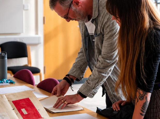 A close-up view of a man and a woman standing over a table to carefully examine historic archival documents and a large vintage folder.