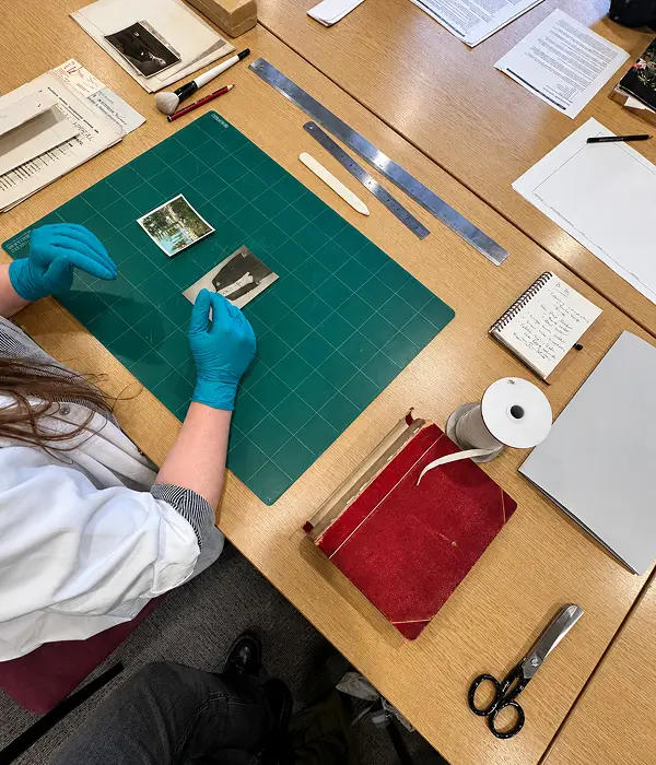 Overhead view of a volunteer wearing blue nitrile gloves examining a historic photograph on a green cutting mat, surrounded by archival tools, documents, a red bound volume, and conservation materials on a wooden worktable.