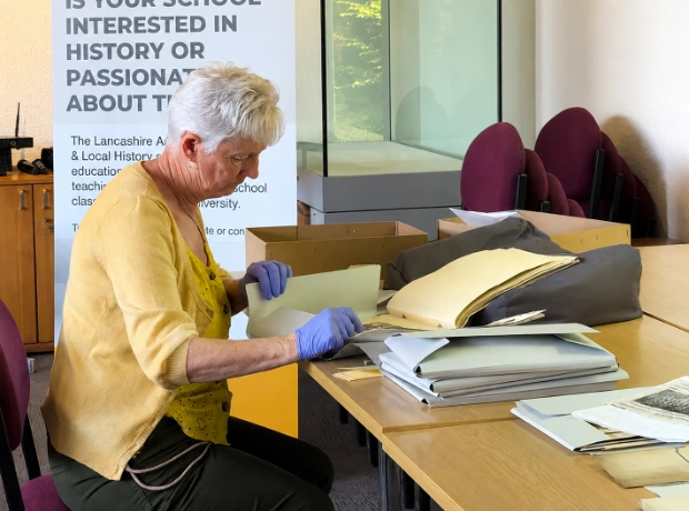 An archivist wearing purple protective gloves sits at a long table, carefully sorting through historical folders and documents at the Lancashire County Council offices.