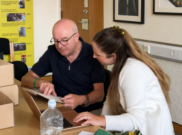 A man and a woman collaborating at a desk, with the man pointing at an open archival book.