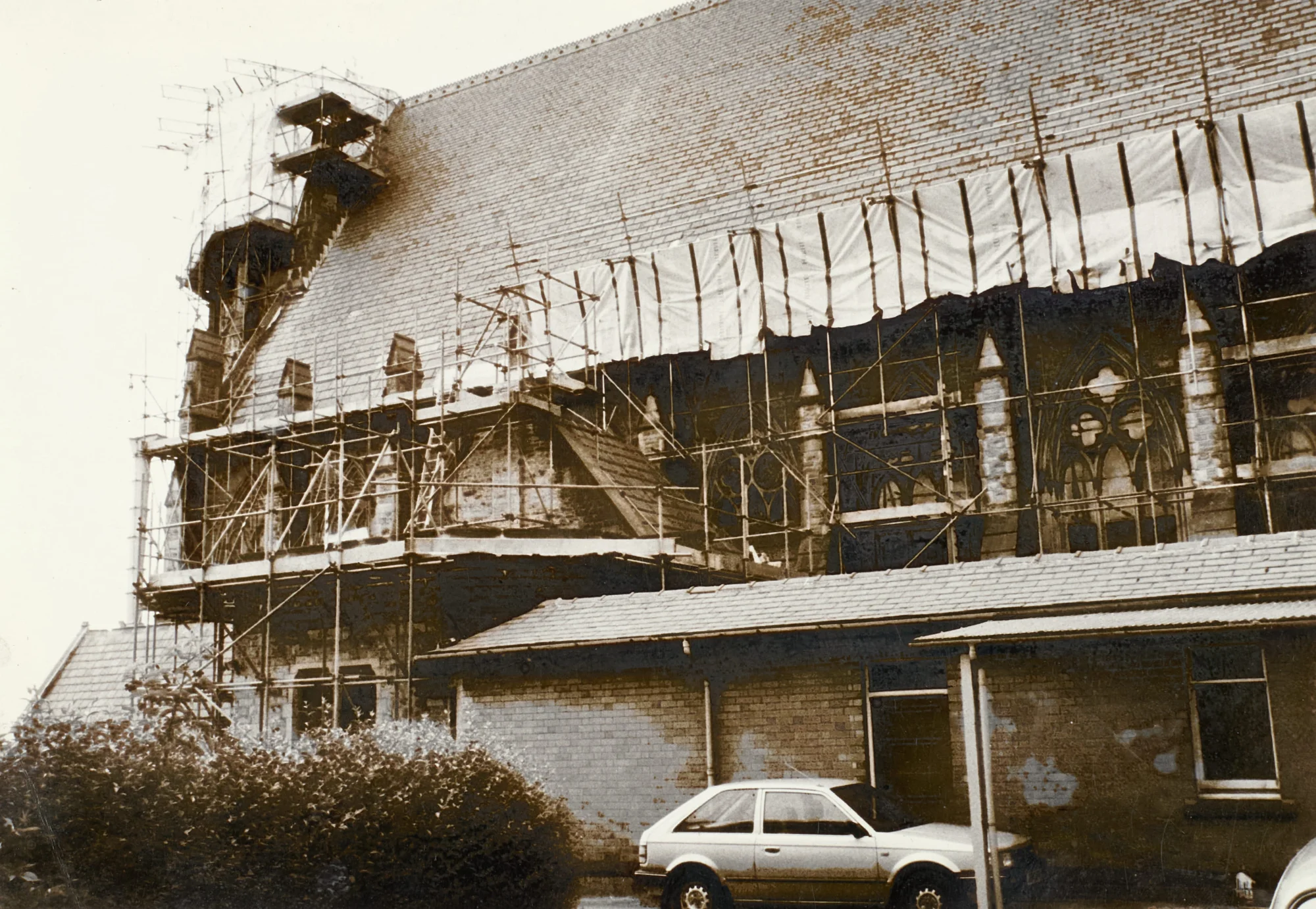 Photograph of St Walburge's exterior during restoration, scaffolding and sheeting rising the full height of the roof and covering the traceried windows below. A car is parked on the road in front, alongside an adjoining parish building.