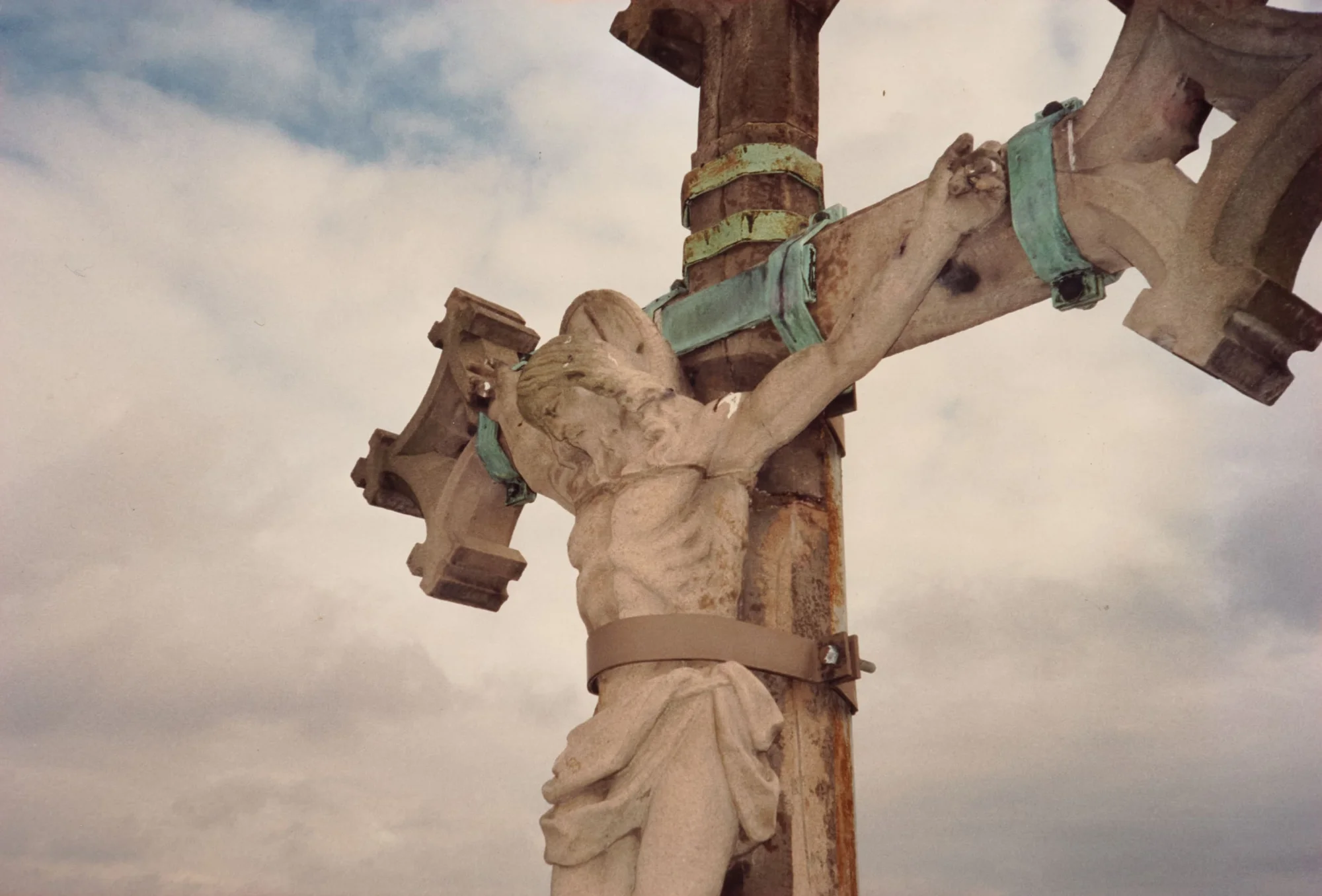 Photograph of the crucifix of St Walburge's Calvary, seen from directly below, the figure held to the cross with metal fixing bands against a clouded sky.
