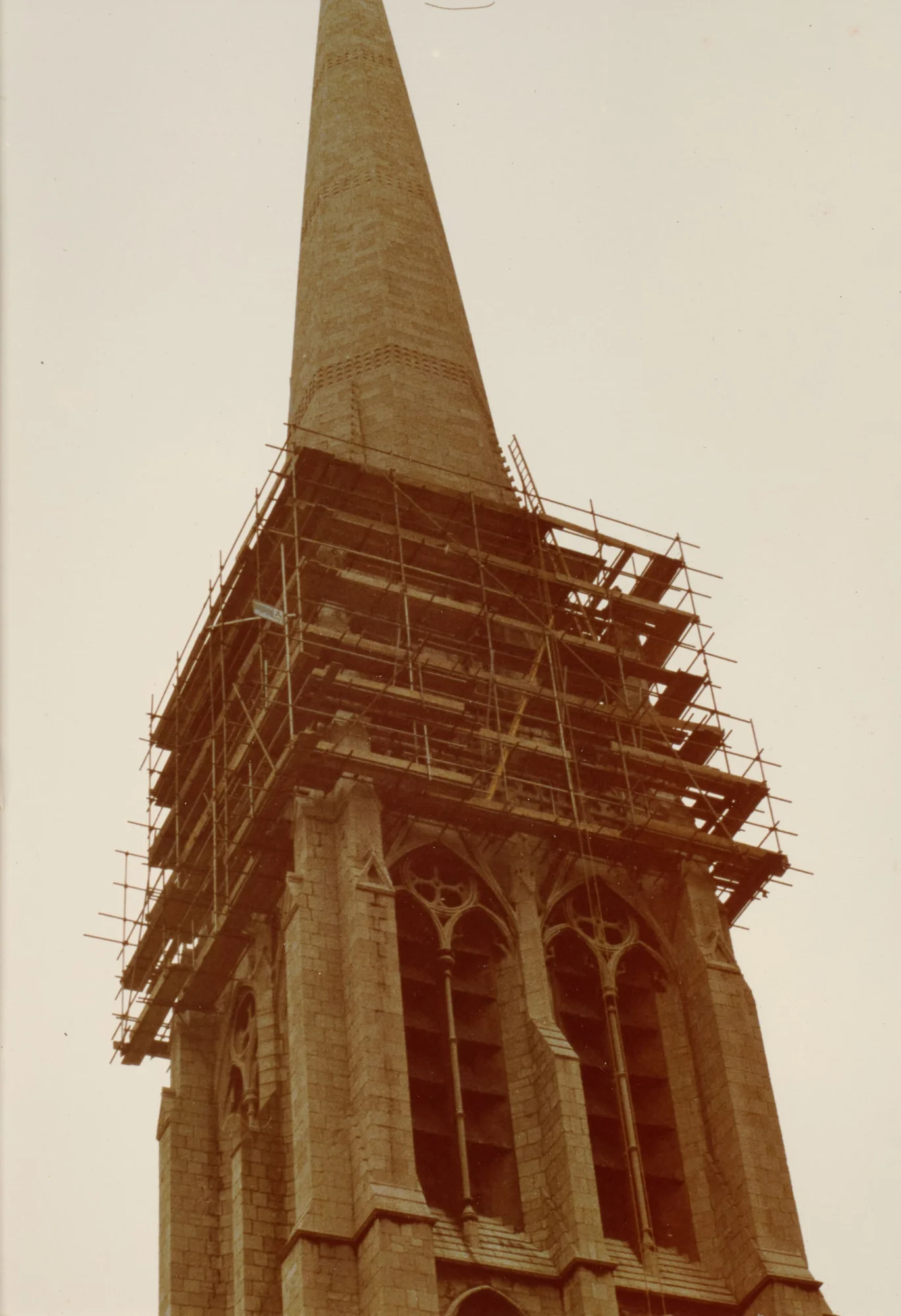 Photograph looking up at St Walburge's spire, scaffolding encircling the tower below the base of the spire while the stonework tapers unobstructed above. Gothic belfry windows are visible in the tower beneath.