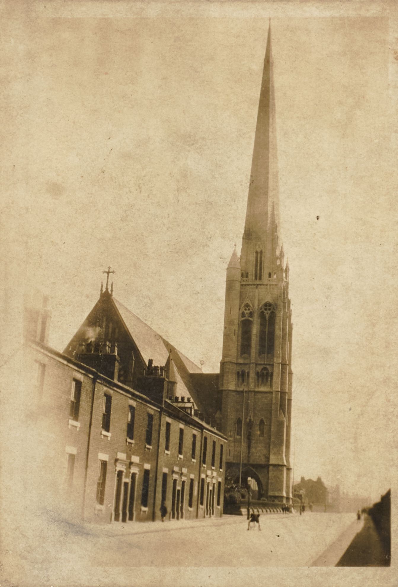 Early photograph of St Walburge's tower and spire beside a row of terraced houses, a figure standing in the empty street below.