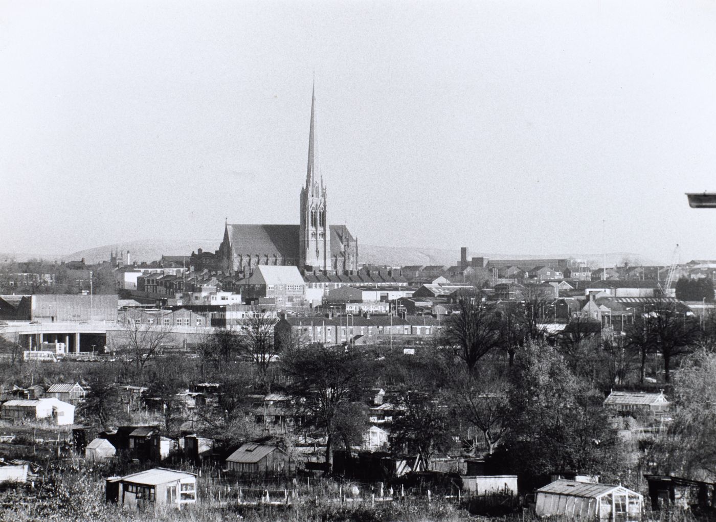 Photograph of St Walburge's spire rising above the town, allotments and greenhouses filling the foreground, a road flyover visible to the left.