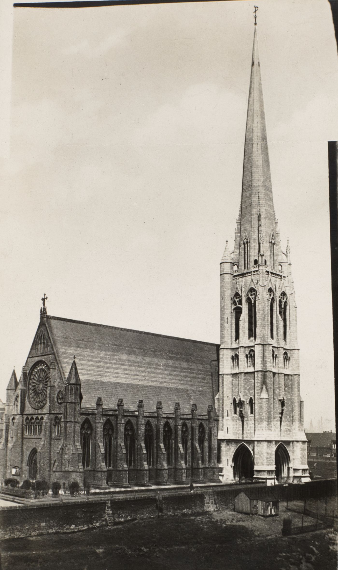 Historical photograph of St Walburge's from a raised vantage, the rose window and gabled west front on the left, the tower and spire rising beside it, open ground in the foreground.