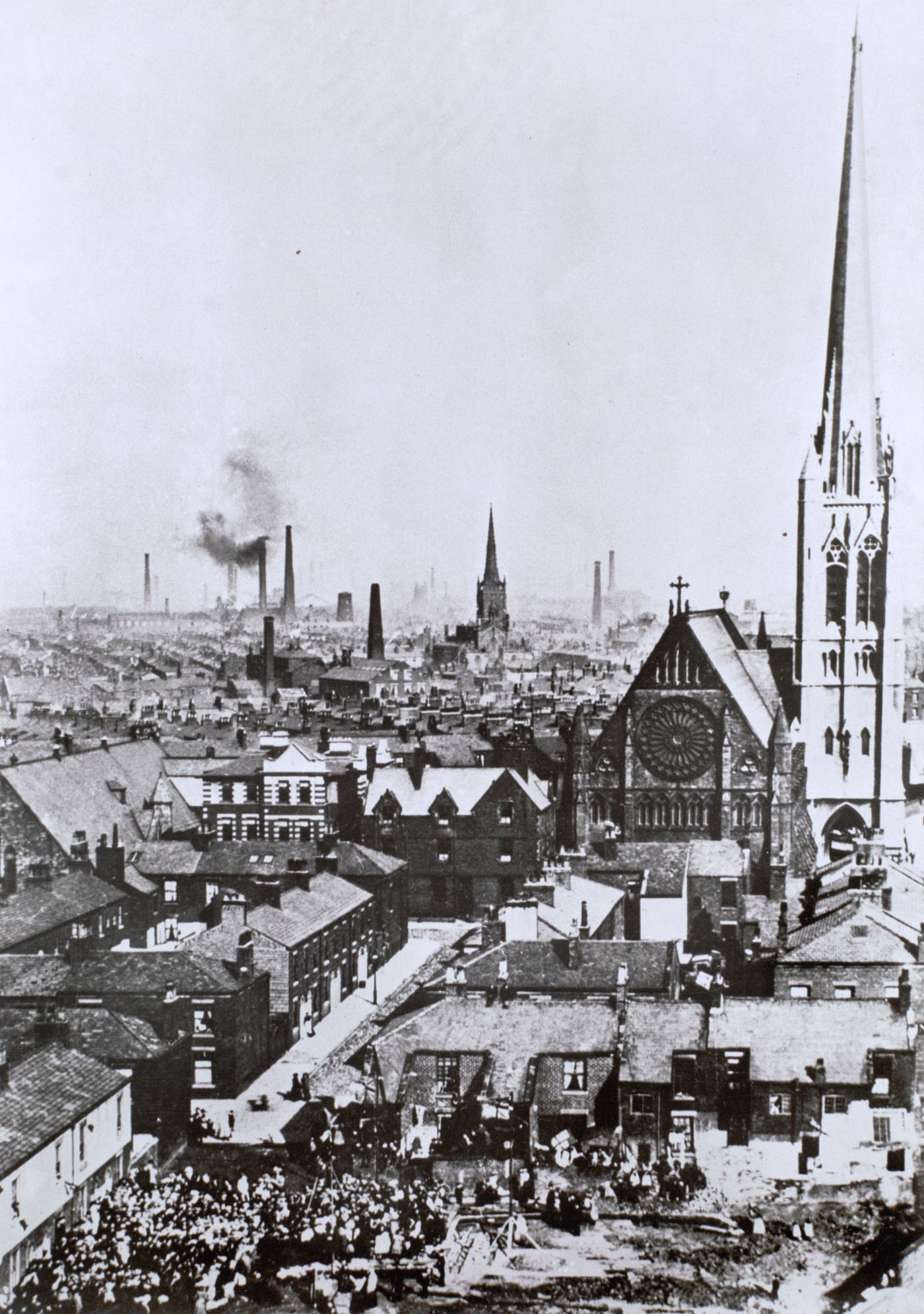 Historical photograph looking over Preston's rooftops and mill chimneys, St Walburge's rose window and spire rising above the terraces, a crowd gathered in the street below.