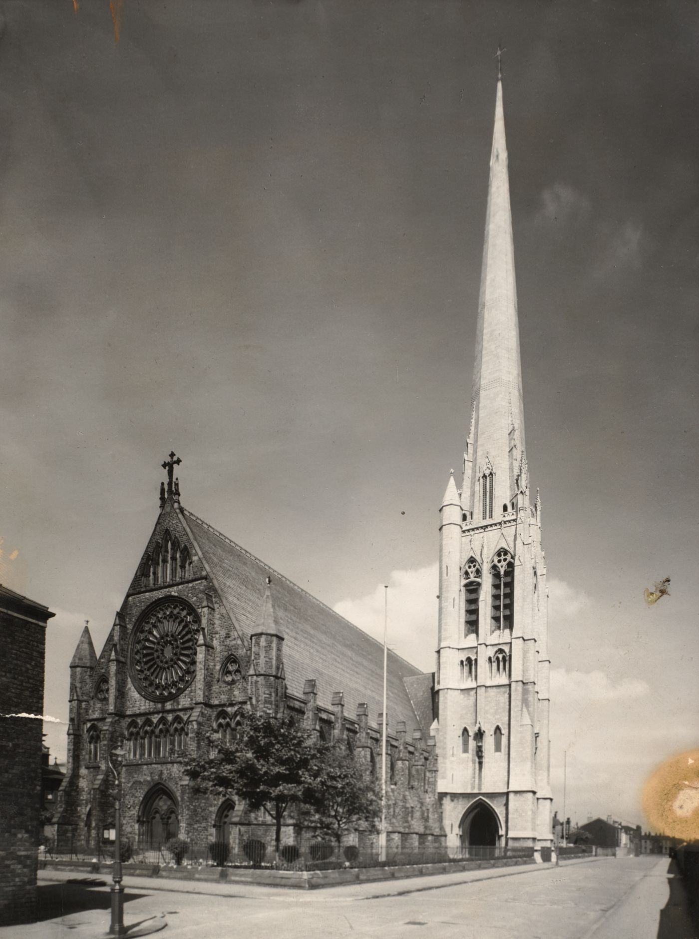 Photograph of St Walburge's from the street, the great rose window and gabled west front on the left, the tall spire rising beside it against a cloudy sky.