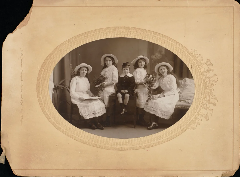 Oval studio portrait of four girls in white dresses and hats, holding flowers, seated with a boy in a sailor-style suit, mounted on a card printed with the photographer's name.
