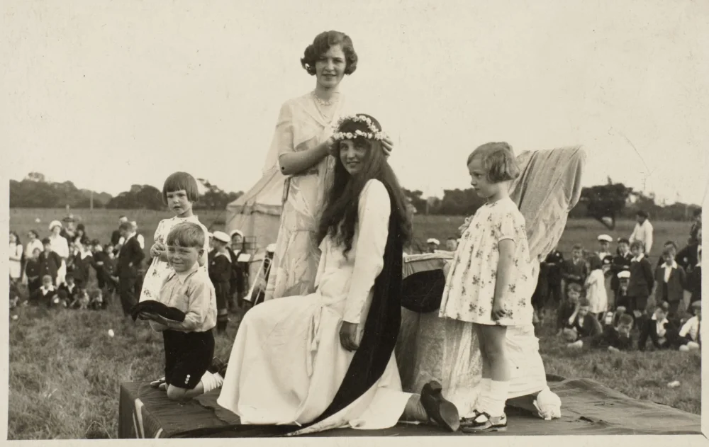 A young woman is crowned with a floral wreath as May Queen, seated on a platform in a field with attendants and a watching crowd behind.