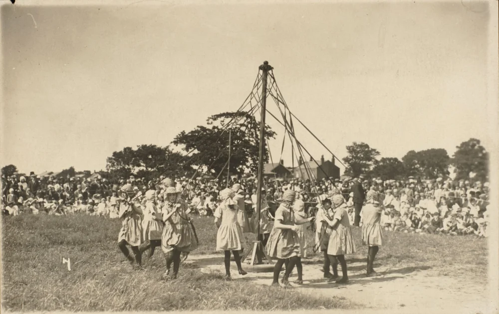 Girls in matching dresses and hats dance around a maypole in a field, watched by a large crowd gathered behind.