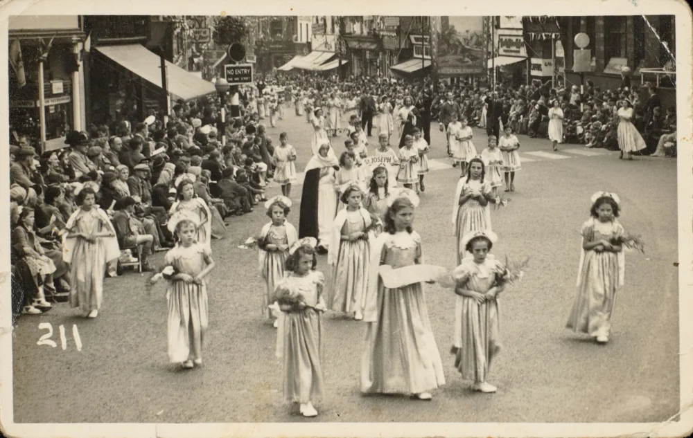 Girls in white processional dress carry flowers along a street lined with spectators, a nun walking behind a "St Joseph" banner.