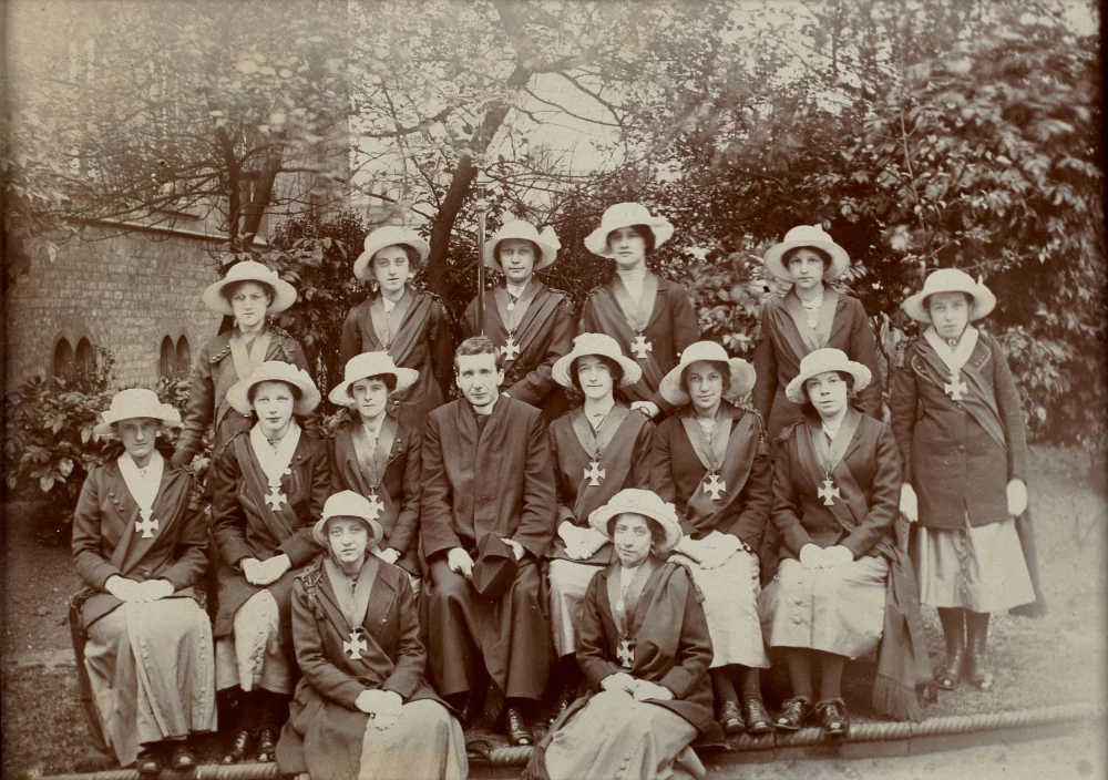 Group photograph of women in matching dark coats, white hats and sashes with cross-shaped badges, a priest seated centrally, in the church grounds.