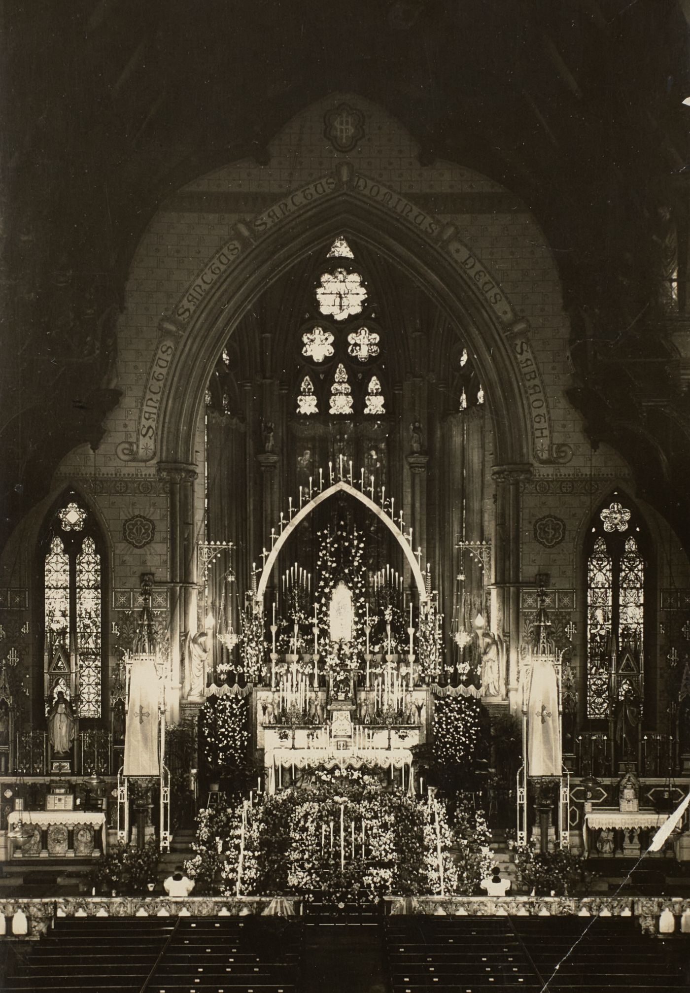 Photograph of St Walburge's sanctuary banked with tiered candles and flowers before the east window, two servers kneeling below.