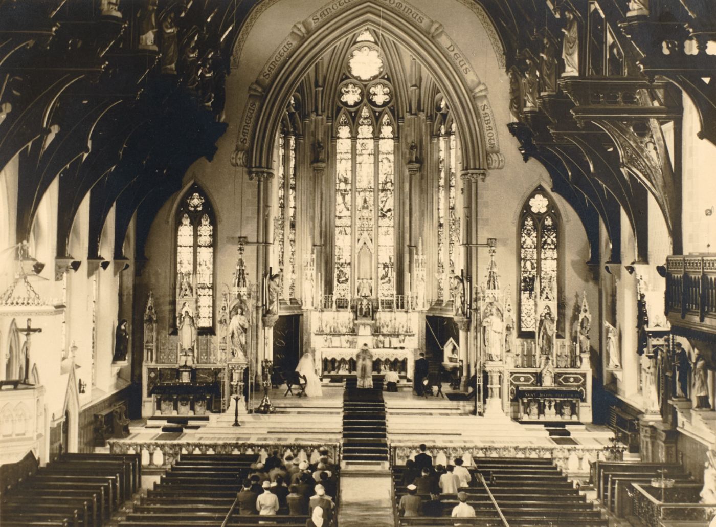 Photograph of St Walburge's sanctuary and east window from the pews, a priest at the altar with a small congregation seated below, side altars and statues either side.