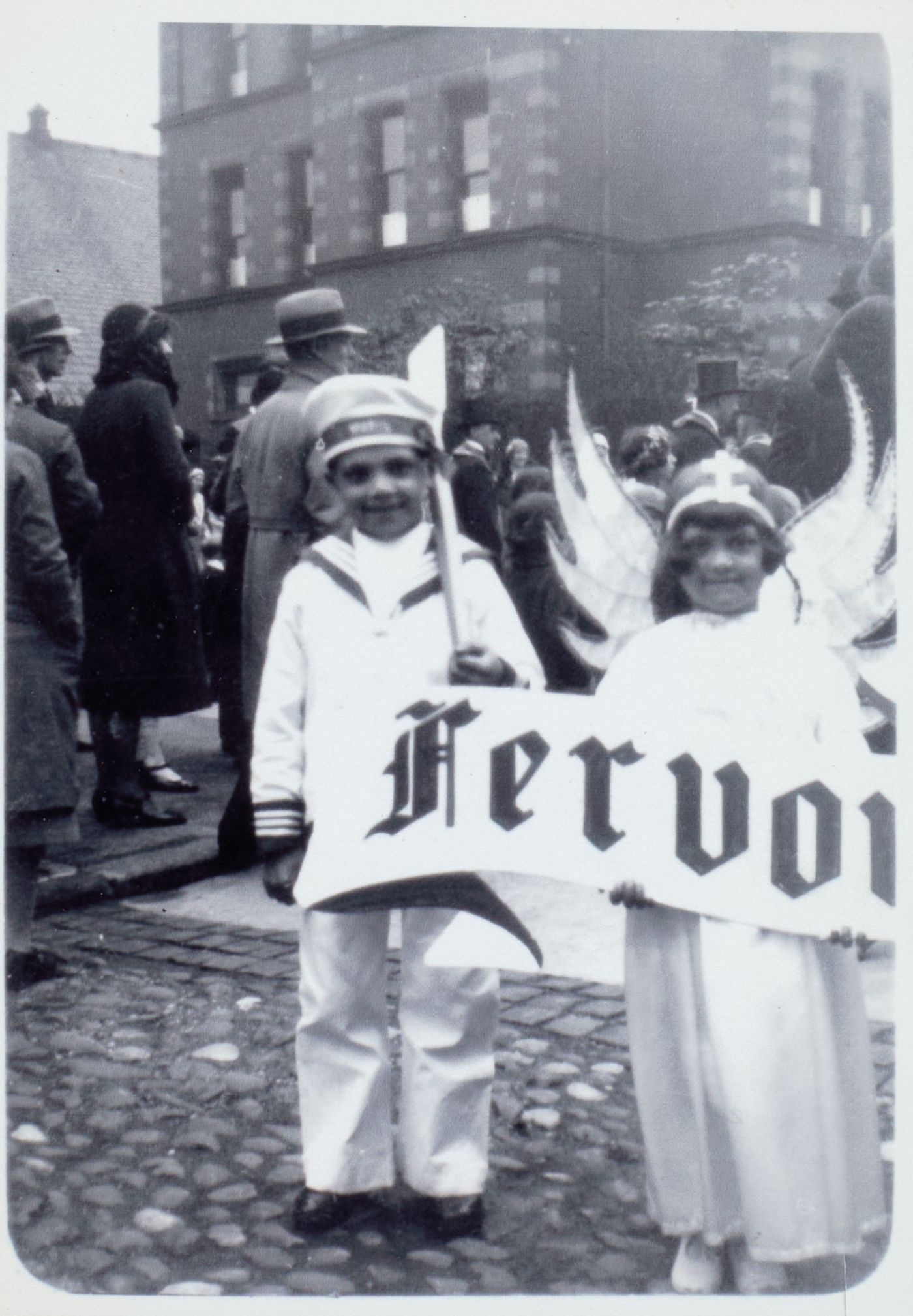 A boy in a sailor suit and a girl dressed as an angel hold a banner reading "Fervour," a crowd gathered behind them in the street.