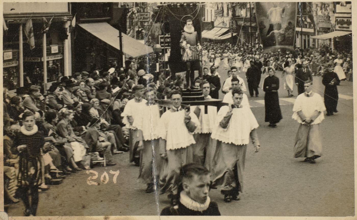 A statue carried on a platform by men in cassock and cotta through a crowded street, crowds seated along the pavement.