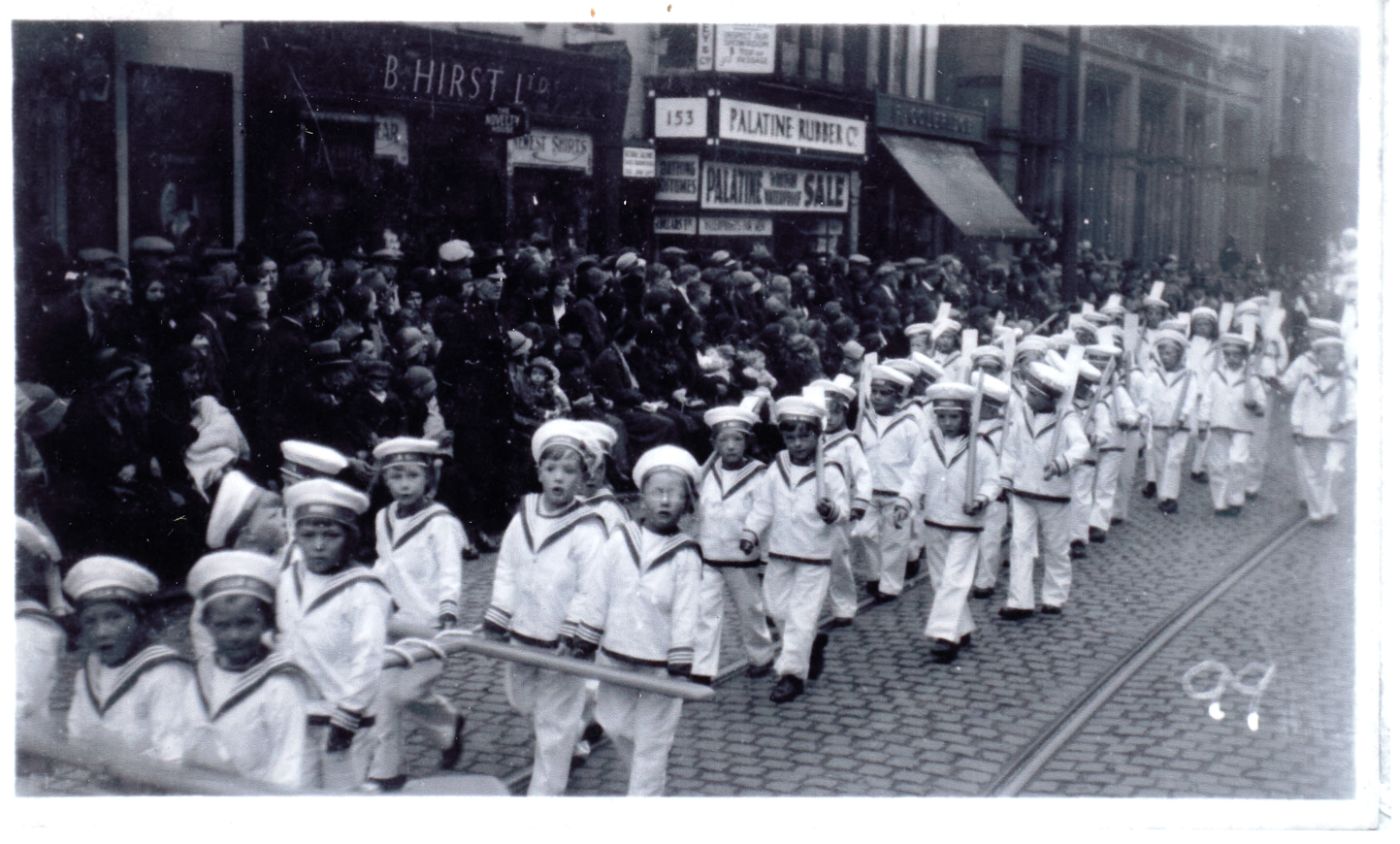 Boys in matching white sailor suits and caps march in rows down a cobbled street, crowds watching outside shopfronts including B. Hirst Ltd and the Palatine Rubber Co.