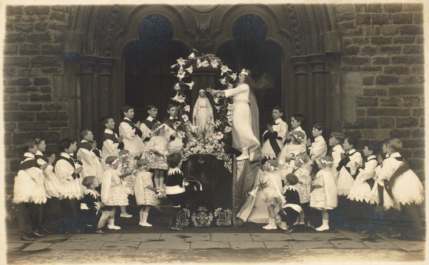 A crowned girl in white reaches to place flowers or a crown at a statue of Our Lady, altar servers and children in white gathered around, at the arched entrance of the church.