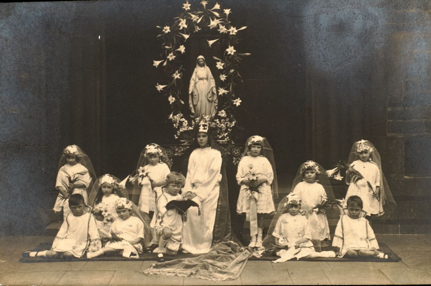 Children in white communion dress and veils, holding lily bouquets, are arranged before a statue of Our Lady wreathed in lilies.