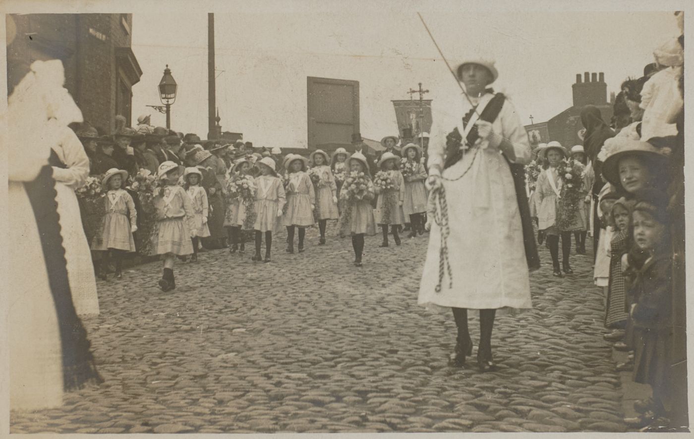 Girls in white dresses and sashes carry flowers along a cobbled street, a woman in white leading with a processional cross behind, crowds watching from the pavement.