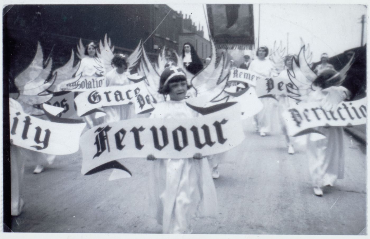 Girls dressed as angels carrying virtue banners including "Fervour" and "Grace," processing down a street.