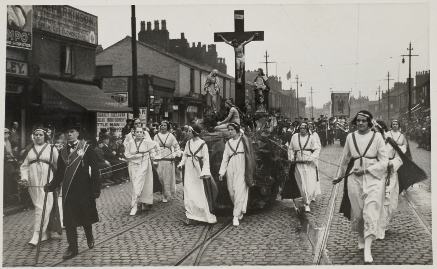 A large Calvary group of the crucified Christ, Our Lady and St John is drawn on a float through a cobbled street, women in white robes and sashes hauling on ropes alongside, crowds watching from the pavement.