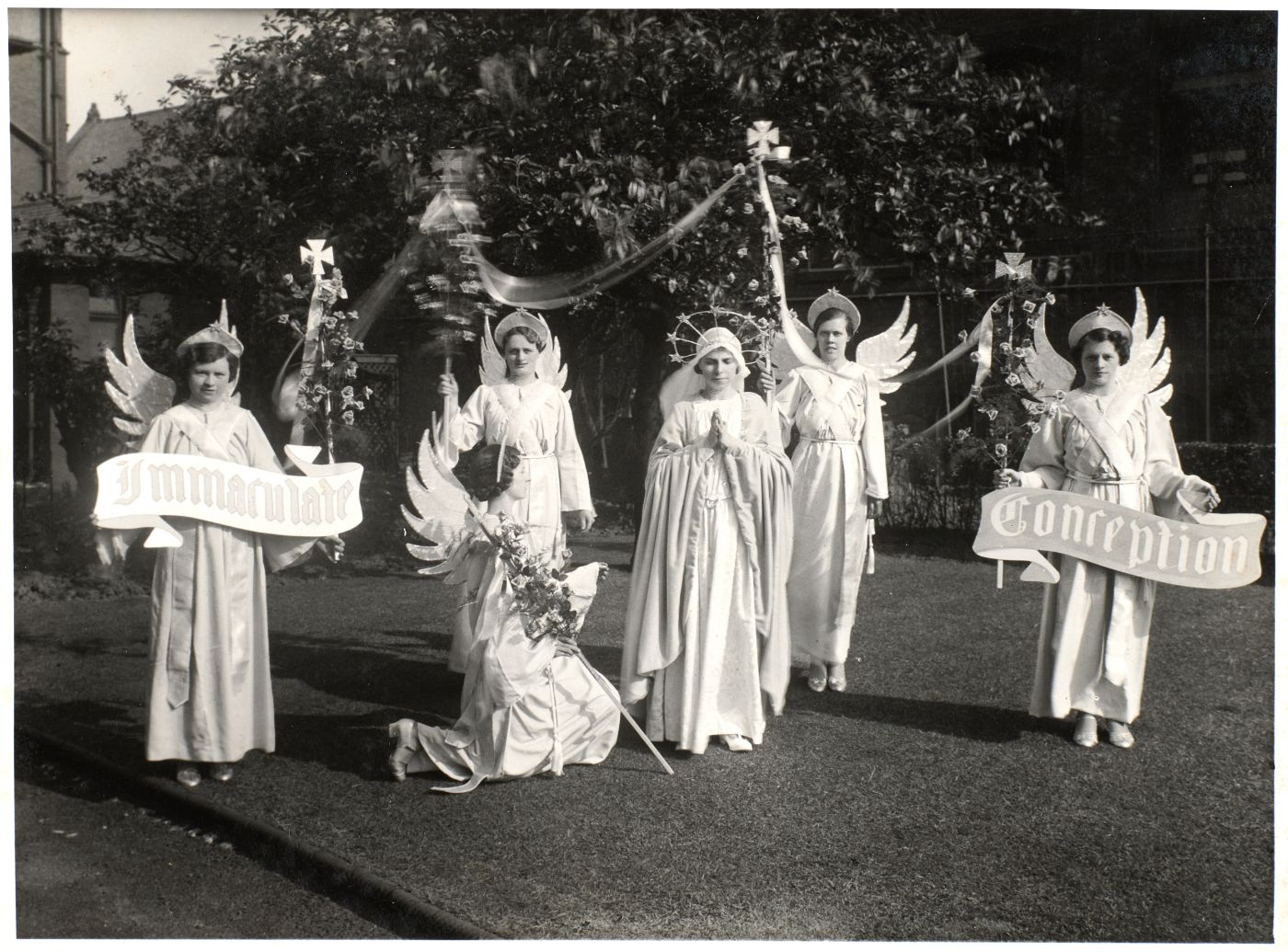 Young women dressed as angels stand around a central figure in a crown and veil with hands joined in prayer, banners reading "Immaculate Conception" held at either end.