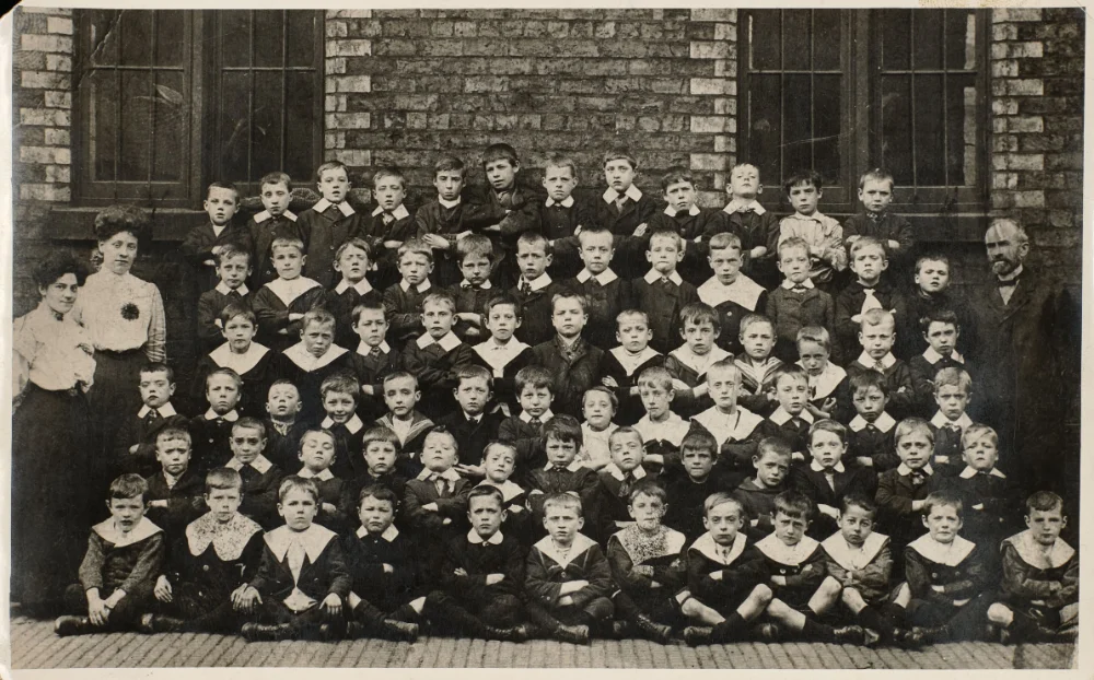 A large class of boys in sailor collars gathers in tiered rows outside the school, with two women and a man standing at the edges.