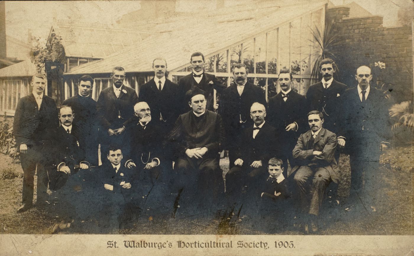 Group photograph of men and boys in front of a glasshouse, a priest seated centrally, captioned "St Walburge's Horticultural Society, 1905."