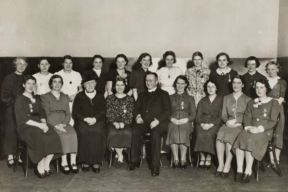 A priest sits among two rows of women, several wearing badges on their coats, gathered indoors for a group photograph.