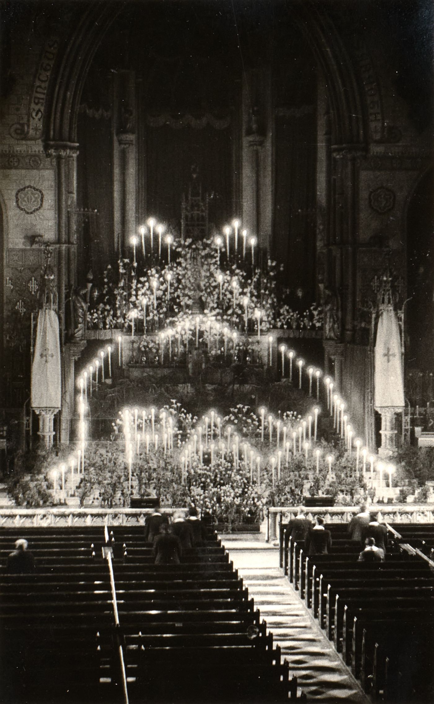 Photograph of St Walburge's sanctuary banked with candles and flowers for the Forty Hours' Devotions, worshippers kneeling in the pews before it.