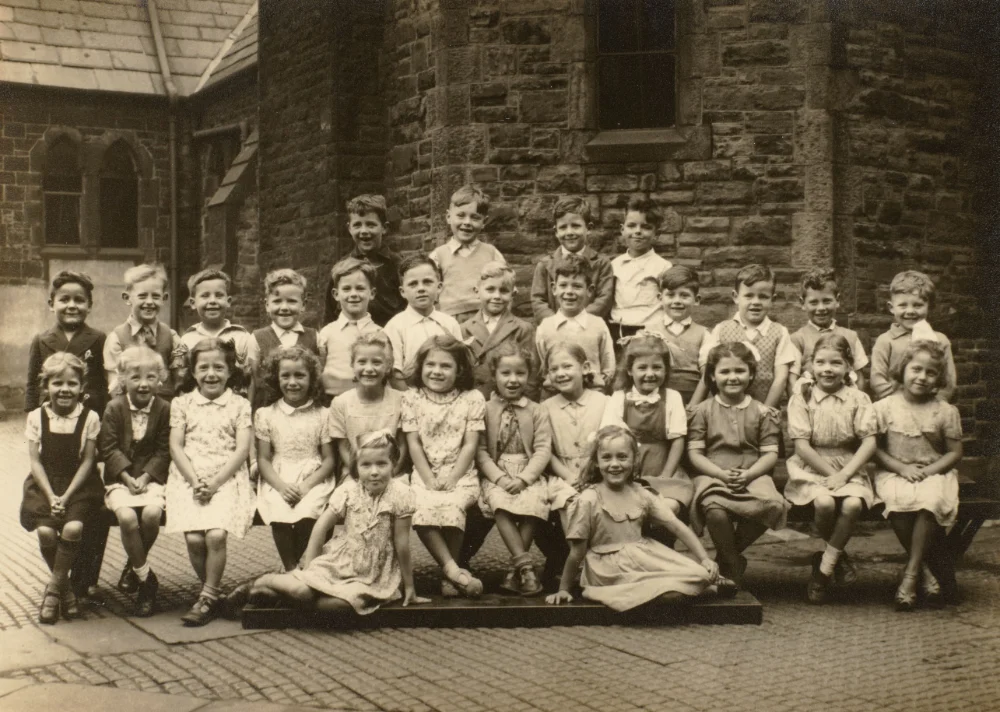 A class of young pupils gathers in tiered rows outside the church for a group photograph, boys standing and girls seated.