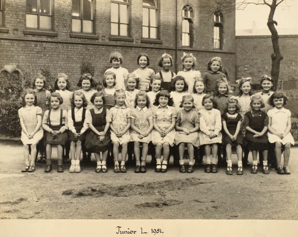 Girls of Junior 1 gather in rows in the schoolyard for a class photograph, captioned "Junior 1 – 1951."