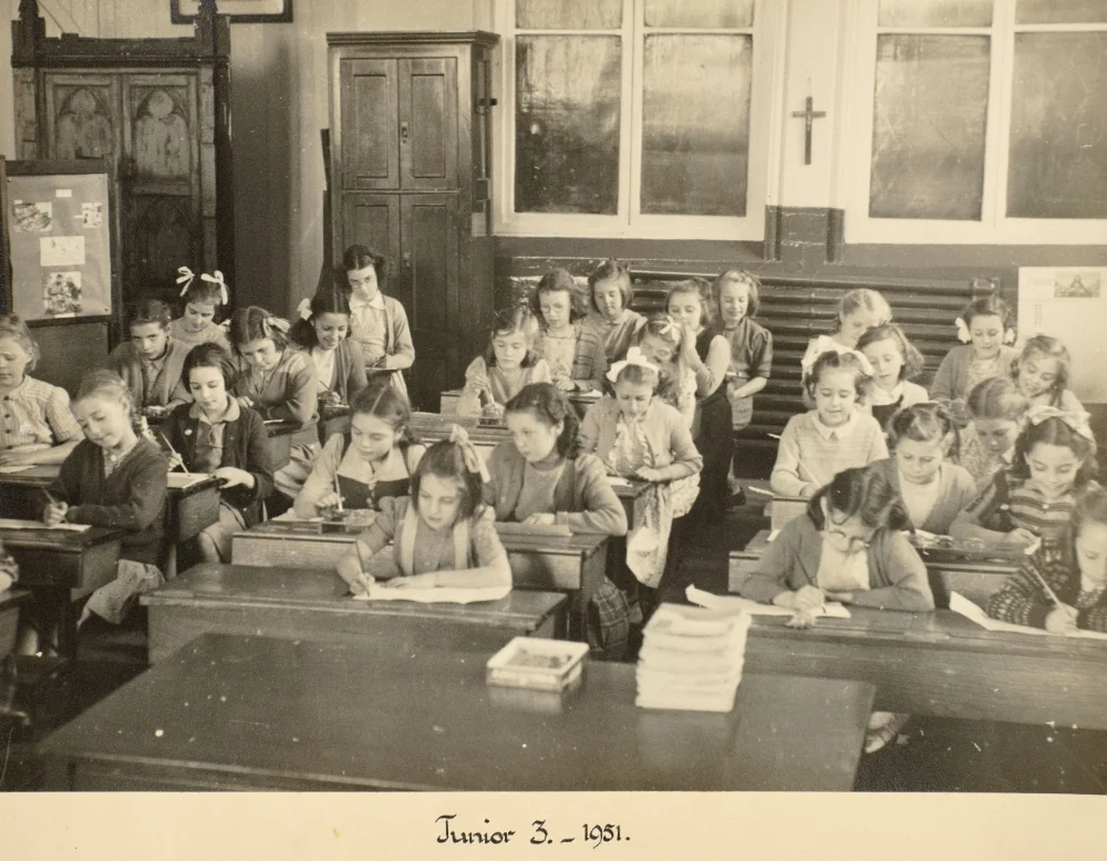 Girls of Junior 3 sit at wooden desks writing, with others standing behind, a wall crucifix visible, captioned "Junior 3 – 1951."